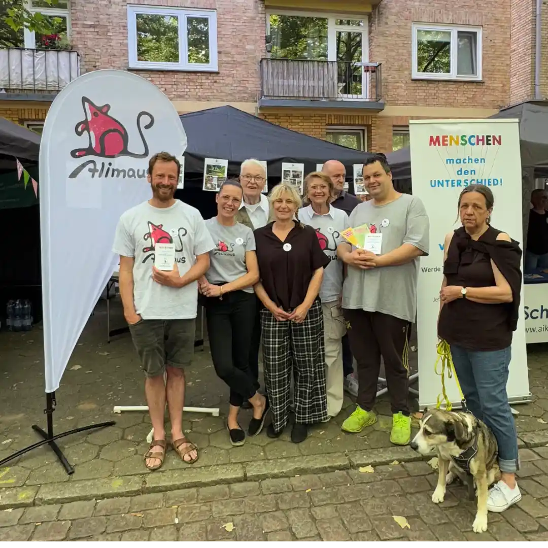 Das Bild zeigt mehrere Menschen, die freundlich in die Kamera schauen. Links steht eine Beschlag mit dem Logo der Alimaus.
