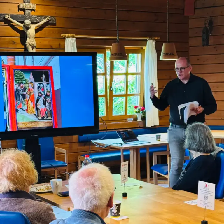 Das Bild zeigt Hauptpastor Dr. Stefan Holtmann, St. Jakobi, bei seinem Vortrag auf der Mitgliederversammlung des Hilfsverins neben einem großen Monitor und darauf eine Präsentation. Im Vordergrund sitzen Teilnehmende der Versammlung und schauen auf den Monitor.
