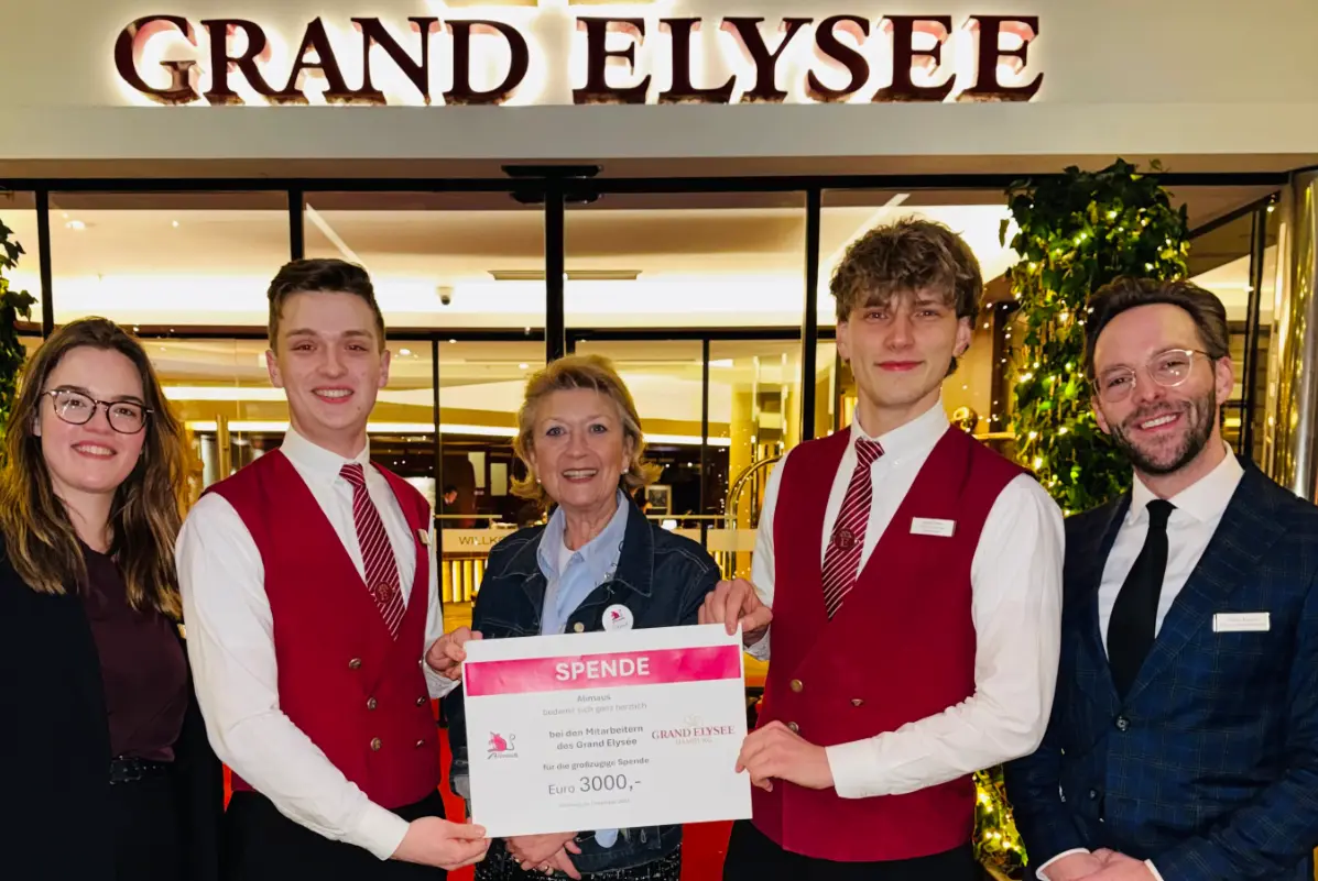 Das Bild zeigt fünf Menschen, die in einer Reihe stehen und die Kamera schauen. Die drei Personen in der Mitte halten einen symbolischen Scheck über 3000 Euro in der Hand. Im Hintergrund ist der Eingang mit dem Schriftzug des Hotels Grand Elysee sichtbar.