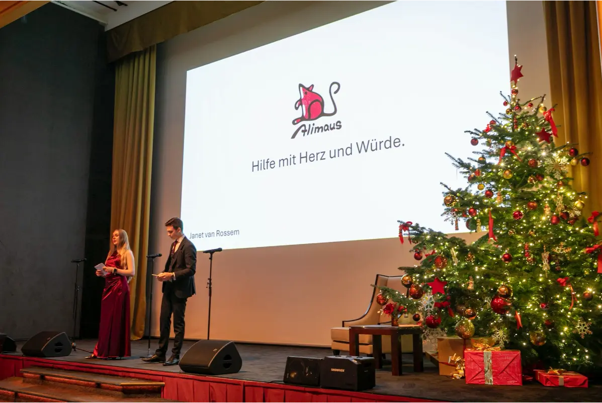 Das Bild zeigt eine Bühne mit Mikrofonen, Lautsprechern, einem geschmückten Weihnachtsbaum mit Geschenkpaketen darunter und im Hintergrund einer Leinwand. Auf der Leinwand ist das Logo der Alimaus sichtbar. Weiterer Text auf der Präsentation: Hilfe mit Herz und Würde. Janet van Rossem". Vorne an den Mikrofonen stehen zwei Personen in festlicher Abendkleidung.